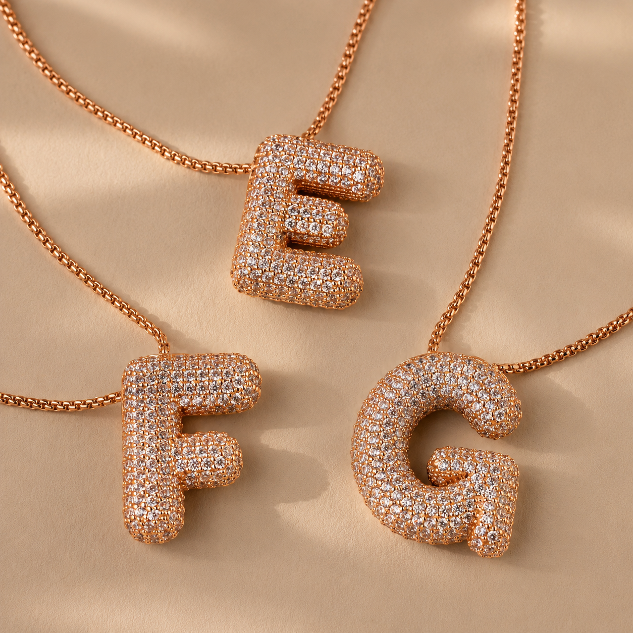 Rose gold letter pendants 'E', 'F', and 'G' on a beige background